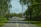 A road with two lanes leads into an urban area with a white car parked near the entrance. Several electric scooters are lined up on the side of the road, and there are various traffic signs and utility poles with wires attached. The area is surrounded by lush green trees and bushes on both sides.