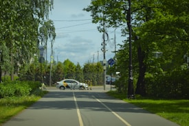 A road with two lanes leads into an urban area with a white car parked near the entrance. Several electric scooters are lined up on the side of the road, and there are various traffic signs and utility poles with wires attached. The area is surrounded by lush green trees and bushes on both sides.