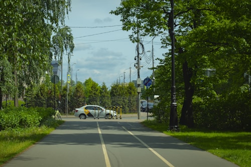 A road with two lanes leads into an urban area with a white car parked near the entrance. Several electric scooters are lined up on the side of the road, and there are various traffic signs and utility poles with wires attached. The area is surrounded by lush green trees and bushes on both sides.