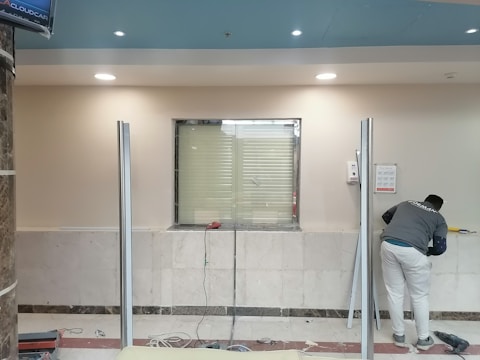 A man is working in a room with a marble-tiled wall and a window surrounded by metal frames. The man is wearing a gray shirt and white pants, using tools for what appears to be an installation or maintenance task. The ceiling is blue with several recessed lights.