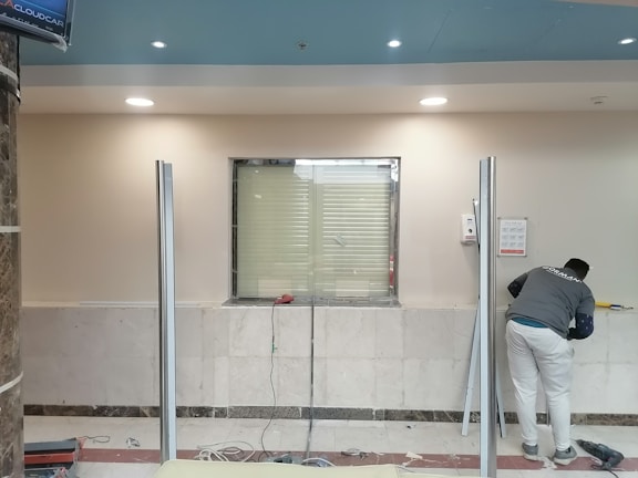 A man is working in a room with a marble-tiled wall and a window surrounded by metal frames. The man is wearing a gray shirt and white pants, using tools for what appears to be an installation or maintenance task. The ceiling is blue with several recessed lights.