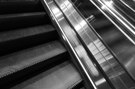 An upward view of escalator steps and their stainless steel side railings, emphasizing a modern, angular design in a monochrome color scheme.