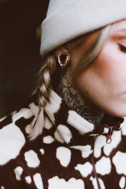 A close-up of a person wearing a light-colored knit beanie and large gold hoop earrings. The individual has a braid and visible tattoo designs on the neck. They are wearing a fleece jacket with a brown and cream abstract pattern.