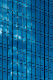 The image features a close-up view of a glass building facade with a grid-like pattern. The glass reflects the blue sky and clouds, creating a dynamic play of light and shadow.