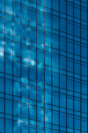 The image features a close-up view of a glass building facade with a grid-like pattern. The glass reflects the blue sky and clouds, creating a dynamic play of light and shadow.