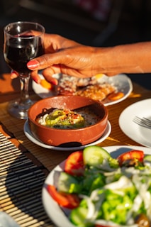 A hand with painted nails is holding a glass of red wine above a table that features a dish of baked cheese topped with pepper slices and herbs, a salad with cucumbers, lettuce, and tomatoes, and a dessert dish. The scene is set outdoors with plenty of natural light.