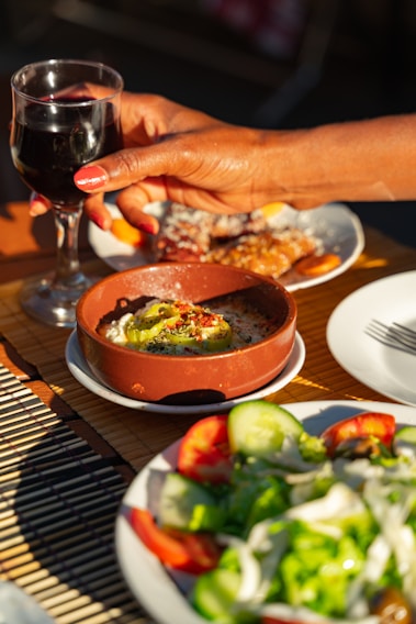 A hand with painted nails is holding a glass of red wine above a table that features a dish of baked cheese topped with pepper slices and herbs, a salad with cucumbers, lettuce, and tomatoes, and a dessert dish. The scene is set outdoors with plenty of natural light.