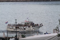 A large passenger boat navigates through calm waters, crowded with people standing and sitting on its deck. It carries a Croatian flag at the stern and a sign on its side reads 'Dubrovnik boat'. Surrounding the water are rocky shores, and white umbrellas are visible in the foreground.
