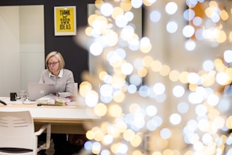 A woman with blonde hair and glasses is seated at an office desk, using a laptop. The desk is organized with a few items, including a black mug and some documents. In the background, there is a motivational poster that reads 'THINK OF YOUR OWN IDEAS' on the wall. The image features a bokeh effect with bright circular lights dominating the right side.
