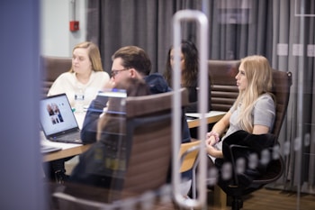 A group of people are seated around a conference table, engaged in a meeting. Some have laptops open in front of them, and papers are scattered on the table. The setting is an office environment with glass walls and brown leather chairs.