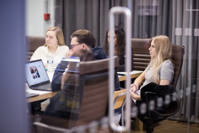 A group of people are seated around a conference table, engaged in a meeting. Some have laptops open in front of them, and papers are scattered on the table. The setting is an office environment with glass walls and brown leather chairs.