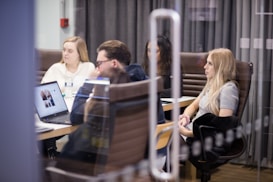 A group of people are seated around a conference table, engaged in a meeting. Some have laptops open in front of them, and papers are scattered on the table. The setting is an office environment with glass walls and brown leather chairs.