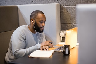 A man is sitting in a quiet, private booth, using a laptop and listening with earphones. There is a notebook and a pen on the table, and a black mug nearby. The setting appears to be a modern workspace with grey cushioned panels and a dark brick wall.