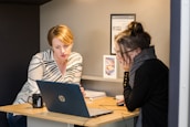 Two women are sitting at a small wooden desk, focusing on a laptop screen. One is wearing a white blouse with black stripes, and the other is in a dark sweater with glasses, resting her face on her hand. There are notebooks, a mug, and some printed materials on the desk.