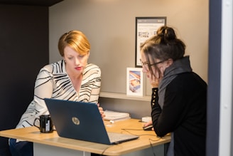 Two women are sitting at a small wooden desk, focusing on a laptop screen. One is wearing a white blouse with black stripes, and the other is in a dark sweater with glasses, resting her face on her hand. There are notebooks, a mug, and some printed materials on the desk.