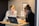 Two women are sitting at a small wooden desk, focusing on a laptop screen. One is wearing a white blouse with black stripes, and the other is in a dark sweater with glasses, resting her face on her hand. There are notebooks, a mug, and some printed materials on the desk.