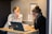 Two women are sitting at a small wooden desk, focusing on a laptop screen. One is wearing a white blouse with black stripes, and the other is in a dark sweater with glasses, resting her face on her hand. There are notebooks, a mug, and some printed materials on the desk.