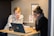 Two women are sitting at a small wooden desk, focusing on a laptop screen. One is wearing a white blouse with black stripes, and the other is in a dark sweater with glasses, resting her face on her hand. There are notebooks, a mug, and some printed materials on the desk.