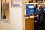 A person sits at a wooden desk working on video editing software seen on dual monitors. They wear headphones and appear focused. The workspace is modern, featuring light brown wooden panels and shelves with decorative items. A mug and keyboard are placed on the desk.