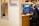 A person sits at a wooden desk working on video editing software seen on dual monitors. They wear headphones and appear focused. The workspace is modern, featuring light brown wooden panels and shelves with decorative items. A mug and keyboard are placed on the desk.