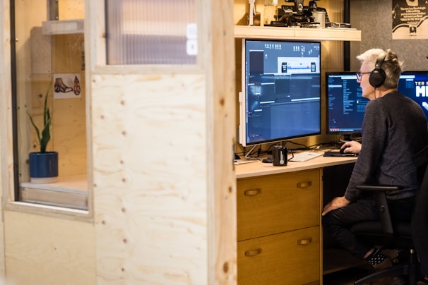 A person sits at a wooden desk working on video editing software seen on dual monitors. They wear headphones and appear focused. The workspace is modern, featuring light brown wooden panels and shelves with decorative items. A mug and keyboard are placed on the desk.