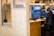 A person sits at a wooden desk working on video editing software seen on dual monitors. They wear headphones and appear focused. The workspace is modern, featuring light brown wooden panels and shelves with decorative items. A mug and keyboard are placed on the desk.