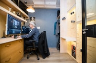A cozy office space with wooden furniture and shelving. A person is seated at a desk working on a computer with large monitors. The room is well-organized, with a coat hanging and camera equipment displayed on shelves.