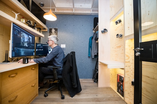 A cozy office space with wooden furniture and shelving. A person is seated at a desk working on a computer with large monitors. The room is well-organized, with a coat hanging and camera equipment displayed on shelves.