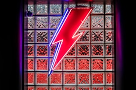 A bright neon lightning bolt in red with a blue outline is set against a decorative glass block wall. The glass blocks have a patterned reflection that amplifies the overall vibrant and electrifying aesthetic.