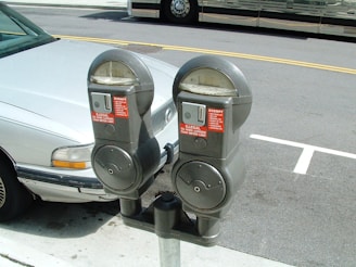 Two gray parking meters are mounted on a single pole. They have labels indicating usage restrictions, situated next to a white car parked on the street.