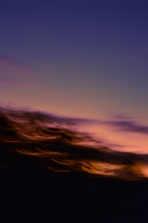 A blurred scene with an abstract blend of colors, featuring a gradient sky transitioning from deep blue at the top to a warm orange and pink near the horizon. The foreground has swirling, motion-like effects in dark shades, giving a dynamic feel.