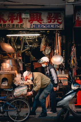 An outdoor street scene in front of a hardware store displays a cluttered assortment of tools and equipment. Two people are present, both wearing masks and hats. One person is working on a bicycle, bending down, while the other stands beside. Various items like pots, barrels, and other metal objects are visible around them.
