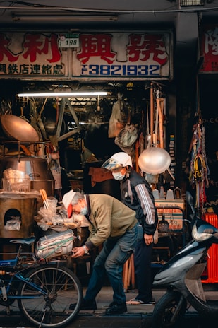 An outdoor street scene in front of a hardware store displays a cluttered assortment of tools and equipment. Two people are present, both wearing masks and hats. One person is working on a bicycle, bending down, while the other stands beside. Various items like pots, barrels, and other metal objects are visible around them.