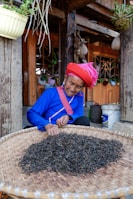 An elderly person wearing traditional clothing with a red headscarf and blue shirt is sorting or inspecting a large quantity of dried herbs or tea leaves spread out on a woven basket. This activity takes place in front of a rustic wooden building with hanging plants and pots, indicating a rural or village setting.