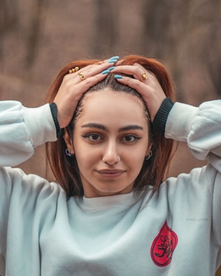 A person with brown hair is holding the top of their head with both hands. They are wearing a white sweater with a red design on the front and have multiple rings on their fingers. The background is blurred trees, suggesting a natural outdoor setting.