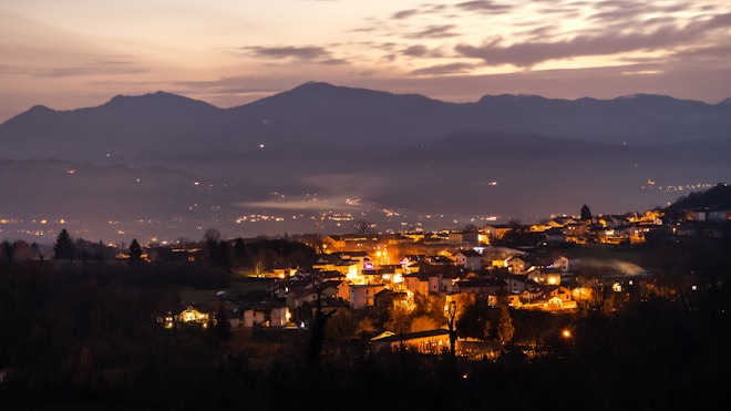 A peaceful village nestled in a valley illuminated by warm, glowing lights at dusk. Rolling hills and distant mountains frame the background, under a sky transitioning from day to night with hues of orange and purple.