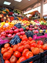 A vibrant and colorful market stall displays a variety of fresh fruits neatly arranged in large quantities. The front showcases bright oranges and pomegranates, with signs displaying their prices. In the background, a diverse assortment of fruits, including mangoes, grapes, and bananas, is visible under the warm indoor lighting.