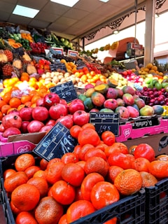 A vibrant and colorful market stall displays a variety of fresh fruits neatly arranged in large quantities. The front showcases bright oranges and pomegranates, with signs displaying their prices. In the background, a diverse assortment of fruits, including mangoes, grapes, and bananas, is visible under the warm indoor lighting.
