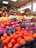 A vibrant and colorful market stall displays a variety of fresh fruits neatly arranged in large quantities. The front showcases bright oranges and pomegranates, with signs displaying their prices. In the background, a diverse assortment of fruits, including mangoes, grapes, and bananas, is visible under the warm indoor lighting.