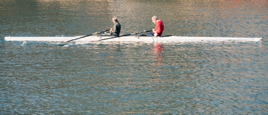 Two people are rowing together in a sleek, narrow boat on a calm body of water. The boat is designed for speed and efficiency, typically used in competitive rowing. The rowers are focused, with one person wearing a red shirt and the other in darker attire.