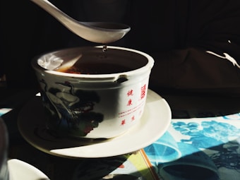 A ceramic bowl with traditional Chinese designs and red characters, filled with a dark liquid. It is placed on a saucer on top of a table with a colorful tablecloth. A spoon hovers above the bowl with a drop of liquid falling into it, illuminated by soft light creating a serene atmosphere.