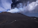 A volcanic landscape with a dark, barren terrain is dominated by a massive plume of smoke and ash erupting from a volcano. The sky above is filled with swirling clouds, contrasting against the dark foreground. A narrow winding road is visible, with small vehicles traveling along it, highlighting the scale of the natural environment.