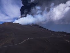 A volcanic landscape with a dark, barren terrain is dominated by a massive plume of smoke and ash erupting from a volcano. The sky above is filled with swirling clouds, contrasting against the dark foreground. A narrow winding road is visible, with small vehicles traveling along it, highlighting the scale of the natural environment.