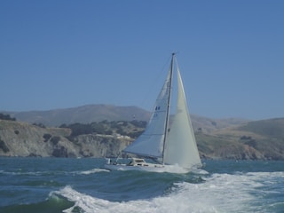A sailboat with white sails gliding on a body of water with waves, against a backdrop of coastal cliffs and rolling hills. The sky is clear with a vibrant blue hue.