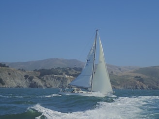 A sailboat with white sails gliding on a body of water with waves, against a backdrop of coastal cliffs and rolling hills. The sky is clear with a vibrant blue hue.