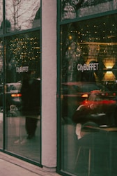 A view of a restaurant named CityBUFFET through large glass windows. The reflections on the glass include holiday lights and passing vehicles, creating a warm and busy atmosphere. Inside, a person is seated, possibly engaged in eating or reading.