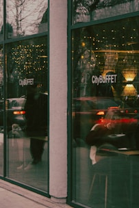 A view of a restaurant named CityBUFFET through large glass windows. The reflections on the glass include holiday lights and passing vehicles, creating a warm and busy atmosphere. Inside, a person is seated, possibly engaged in eating or reading.