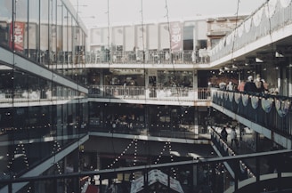 A multi-level shopping complex with open corridors and glass walls. Shoppers are walking along the balconies, and festive lights are strung across the walkways. The architecture features modern elements with metal railings and large windows, while banners hang from the rails promoting sales.