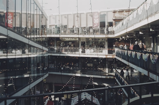 A multi-level shopping complex with open corridors and glass walls. Shoppers are walking along the balconies, and festive lights are strung across the walkways. The architecture features modern elements with metal railings and large windows, while banners hang from the rails promoting sales.