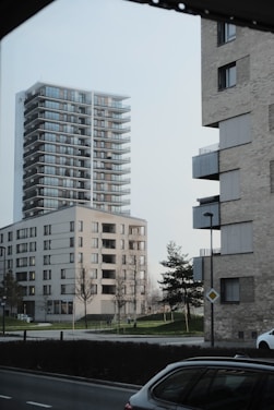 Tall modern residential buildings are situated next to a street in an urban setting. The buildings have multiple balconies and large windows, surrounded by a grassy area with sparse trees. A car is parked on the road, and street signs and lamp posts are present.